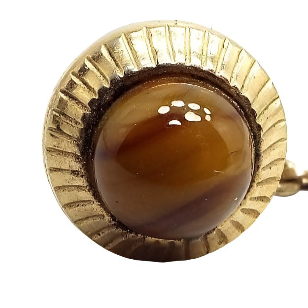 Front view of the Mid Century vintage tie tack. The metal is gold tone in color. It has a round shape with textured lines all the way around the edge. The middle area has a rounded glass cab that has marbled brown and yellow tones. The back clutch has a chain with a small bar on the end.