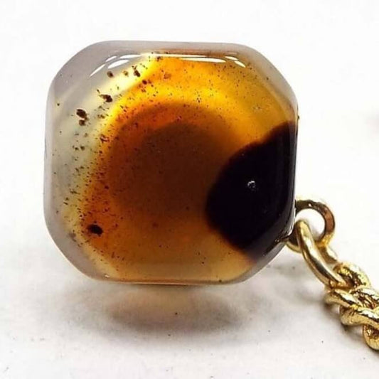 Tie tack with an octagon front that looks like a square with faceted corners. Most of the outer edge is clear in color. In the middle is a round area with shades of orange. On the left side are specks of black and brown color and on the right side is a large splotch of dark brown and black color. There is a gold color chain coming off the back clutch with a bar at the end.