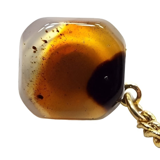Tie tack with an octagon front that looks like a square with faceted corners. Most of the outer edge is clear in color. In the middle is a round area with shades of orange. On the left side are specks of black and brown color and on the right side is a large splotch of dark brown and black color. There is a gold color chain coming off the back clutch with a bar at the end.