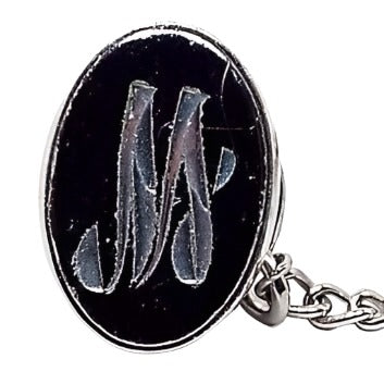 Front view of the Mid Century vintage faux hematite tie tack. The metal is silver tone in color. The tie tack is oval and has a dark metallic gray imitation hemtatite front with a fancy script initial M in the middle. The back clutch has a chain with a bar on the end.