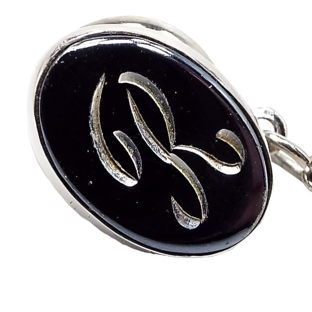 Front view of the Mid Century vintage faux hematite tie tack. The metal is silver tone in color. The tie tack is oval and has a dark metallic gray imitation hemtatite front with a fancy script initial R in the middle. The back clutch has a chain with a bar on the end.