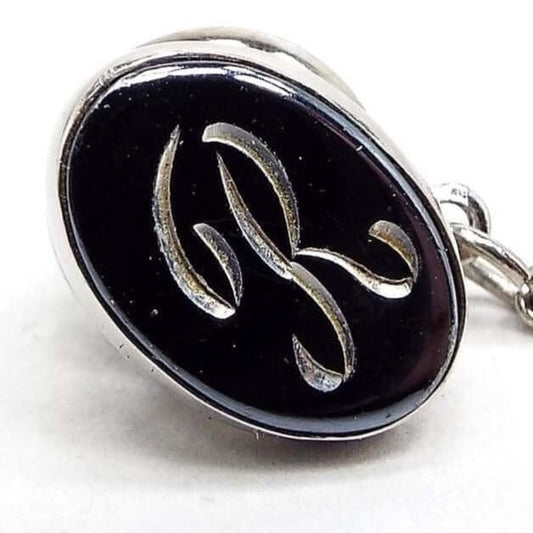 Front view of the Mid Century vintage faux hematite tie tack. The metal is silver tone in color. The tie tack is oval and has a dark metallic gray imitation hemtatite front with a fancy script initial R in the middle. The back clutch has a chain with a bar on the end.
