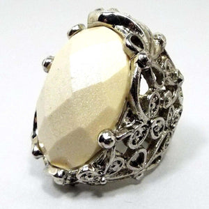 Angled front and side view of the Mid Century vintage chunky large statement ring. The setting and band is silver tone in color. Most of the band has a wide filigree clover like design. The top has a large faceted ivory off white color lucite plastic cab that is prong set. 