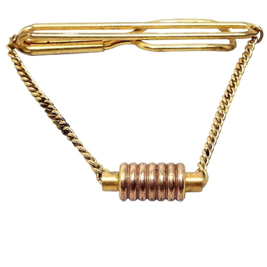 Front view of the 1930's modernist vintage Swank tie bar chain. The metal is gold tone in color. There is a long open oval wire bar at the top that curves around to the back with another open oval area at the end. Curb chain comes down from either side to a charm at the bottom. A few areas of the chain are darkened from age. The charm has a round bar with a coil like design around the middle. 