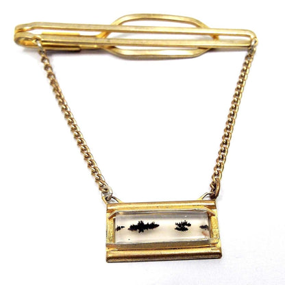 Front view of the 1940's Mid Century Nu Lok vintage tie bar chain.  It has gold tone color metal. The bar at the top is a long open oval. It curves around to the back to form a larger open oval for a slide on style. Curb chain hangs from each side down to a rectangle charm that has a clear and black dendric agate gemstone slice in the middle. Most of the stone is clear with black splotch like areas going like a dotted line across the middle of the gemstone.