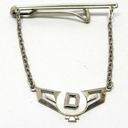 Front view of the 1930's Art Deco vintage tie chain bar. The metal is silver tone in color. It has a long thin double bar at the top with an open wider bar on the back to slide the tie in between. Cable chain runs down from each side of the tie bar down to the letter charm. It has a cut out wide geometric chevron style shape with a round disc of mother of pearl shell on the top. In the middle of the mother of pearl is a silver tone block initial D.