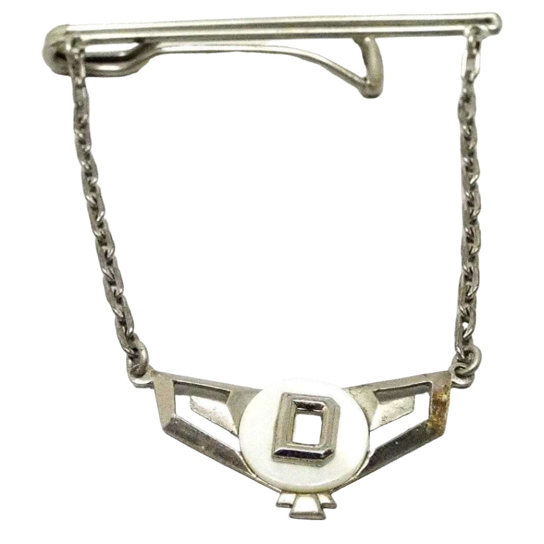 Front view of the 1930's Art Deco vintage tie chain bar. The metal is silver tone in color. It has a long thin double bar at the top with an open wider bar on the back to slide the tie in between. Cable chain runs down from each side of the tie bar down to the letter charm. It has a cut out wide geometric chevron style shape with a round disc of mother of pearl shell on the top. In the middle of the mother of pearl is a silver tone block initial D.