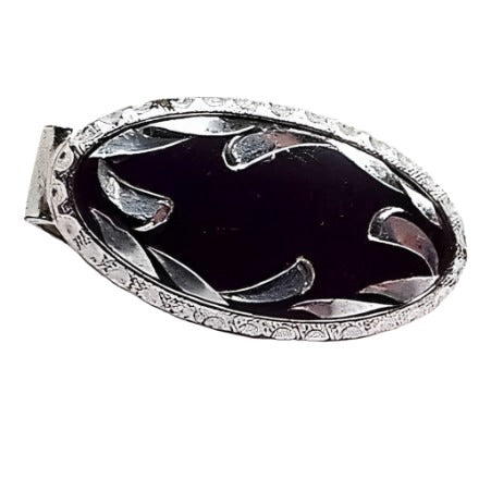 Front view of the Anson Mid Century vintage tie clip. It has a small oval shape with silver tone color metal. The inside of the oval is black with an etched design of curved teardrops flaring inwards. The outer edge of the oval is faceted with a half circle design all the way around.
