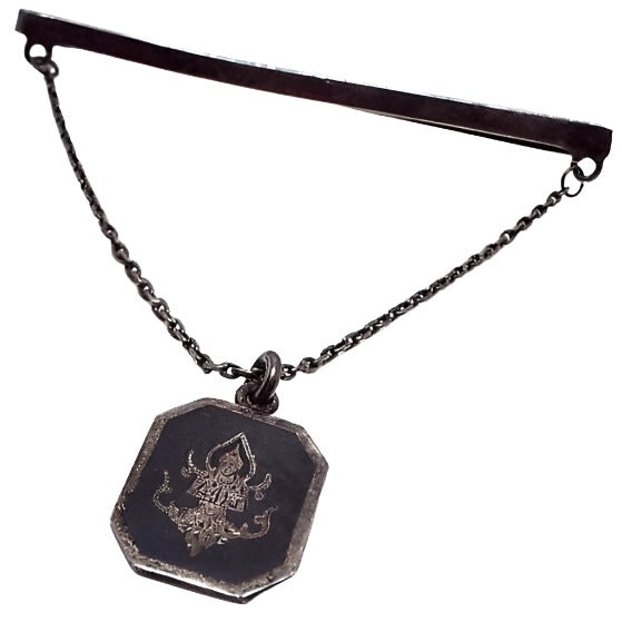 Front view of the enameled sterling silver Mid Century vintage Siam tie chain bar. The sterling is very darkened from age for a dark gray color. The top bar is thin and long and curves around to the back to slide on the tie. There is a thin cable chain coming down from each side to an octagon shaped charm. The charm has an engraved depiction of Thepanom with a black enameled background.