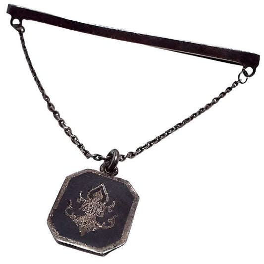 Front view of the enameled sterling silver Mid Century vintage Siam tie chain bar. The sterling is very darkened from age for a dark gray color. The top bar is thin and long and curves around to the back to slide on the tie. There is a thin cable chain coming down from each side to an octagon shaped charm. The charm has an engraved depiction of Thepanom with a black enameled background.