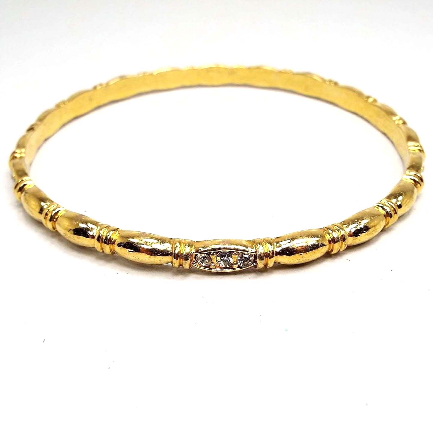 Angled front and side view of the retro vintage rhinestone bangle bracelet. It is gold tone in color with a rounded oval and line design all the way around the edge. There is a small cluster of tiny clear round rhinestones on each side of the bracelet.