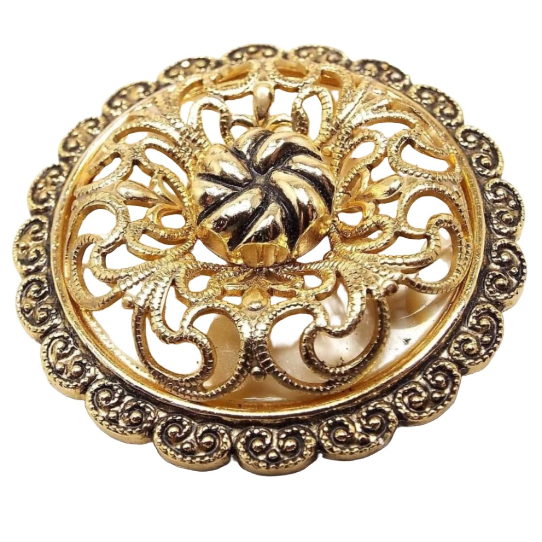 Front view of the Mid Century vintage German scarf clip. It is round in shape with a scalloped textured edge. The top is domed with a filigree design in gold color over a plastic pearly off white background underneath. The middle has a round gold tone bead with a textured twist like appearance. 