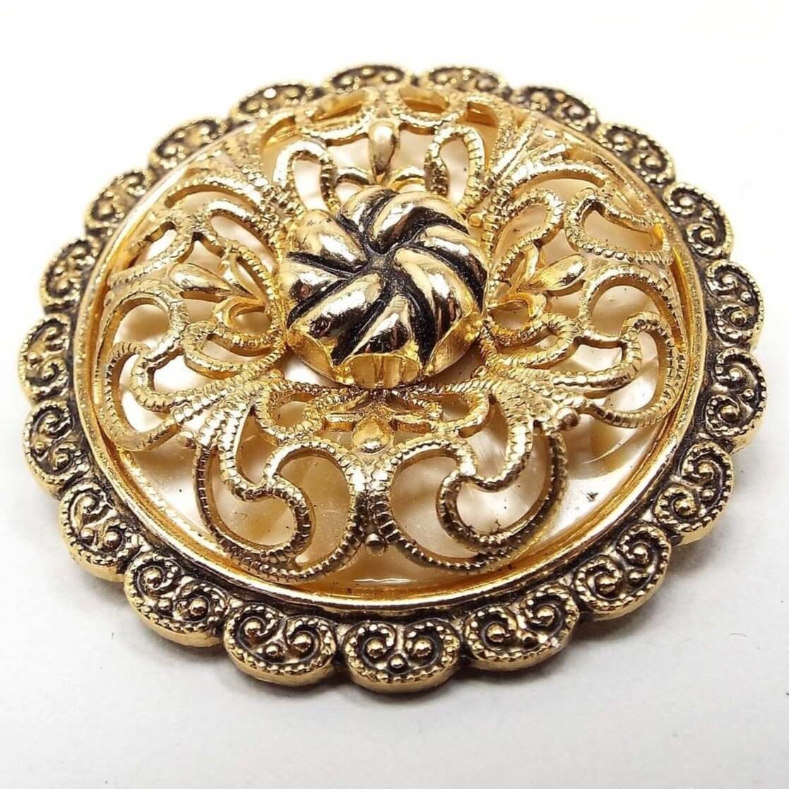 Front view of the Mid Century vintage German scarf clip. It is round in shape with a scalloped textured edge. The top is domed with a filigree design in gold color over a plastic pearly off white background underneath. The middle has a round gold tone bead with a textured twist like appearance. 