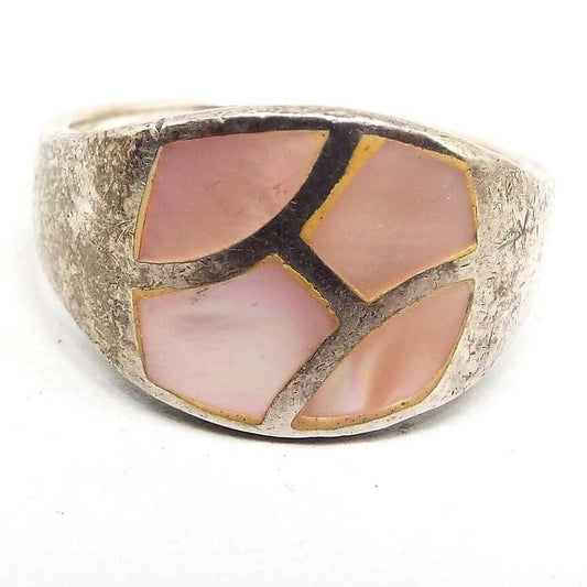 Angled front view of the retro vintage sterling silver mother of pearl band ring. The top part of the ring is wider than the rest of the band with tapered sides. It has a rounded rectangular area that is divided by strips of sterling silver creating four abstract shaped areas of inlaid mother of pearl shell. The shell pieces are dyed a light pink in color. The silver on the rest of the ring has darkened from age.
