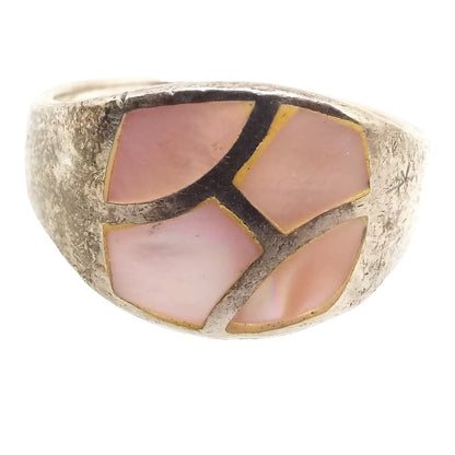Angled front view of the retro vintage sterling silver mother of pearl band ring. The top part of the ring is wider than the rest of the band with tapered sides. It has a rounded rectangular area that is divided by strips of sterling silver creating four abstract shaped areas of inlaid mother of pearl shell. The shell pieces are dyed a light pink in color. The silver on the rest of the ring has darkened from age.