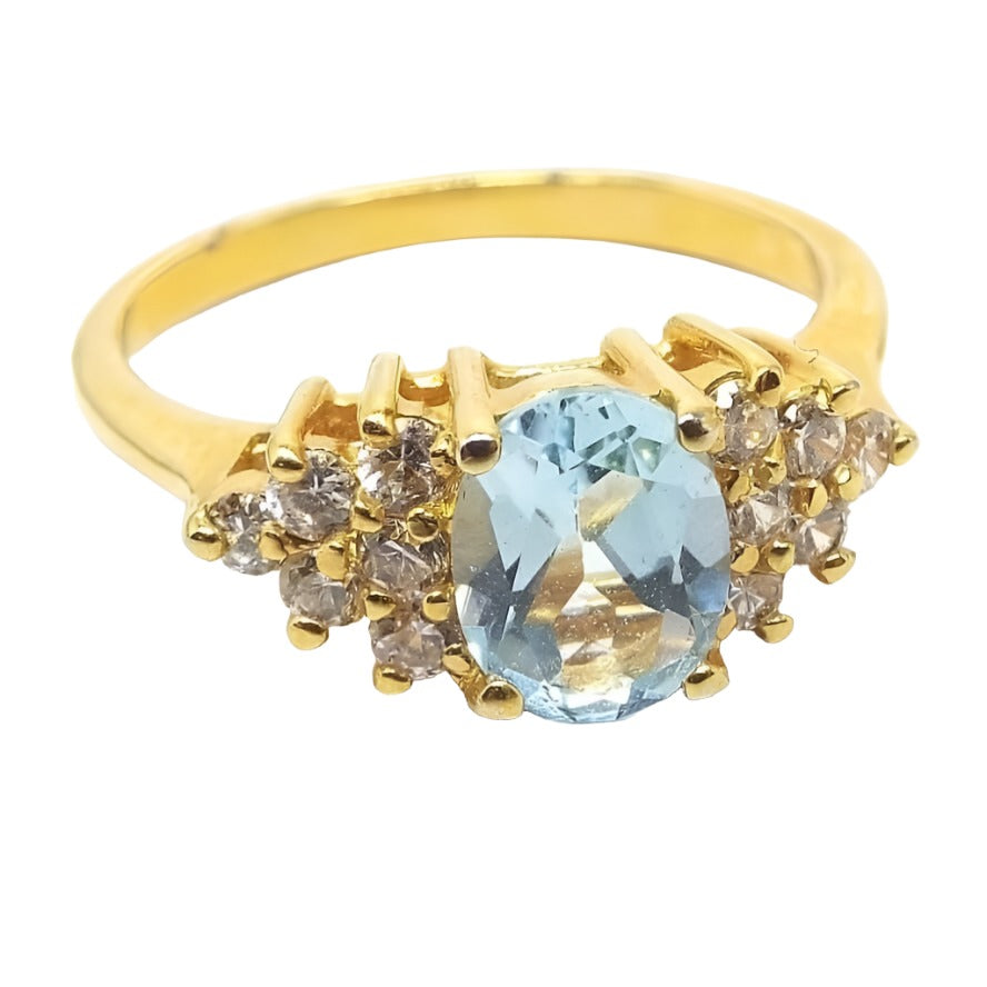 Top view of the retro vintage topaz and cubic zirconia ring. Metal is plated gold tone in color. There is an oval topaz gemstone in the middle that is a very light blue in color. On each side there is a triangle area pointing outwards of small round clear cubic zirconia stones. There are 6 CZ stones on each side. All of the stones are prong set. The band is slightly rounded on the outside and flat and smooth on the inside.