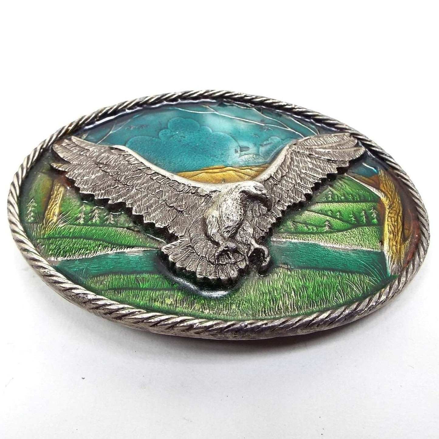Front view of the retro vintage belt buckle from The Great American Buckle Company. The buckle is a gray pewter oval with braided edge. The design is detailed and textured with a flying eagle with its wings spread in the middle. The background is an enameled scenery of grass, trees, and sky, in shades of blue, green, and brown. 