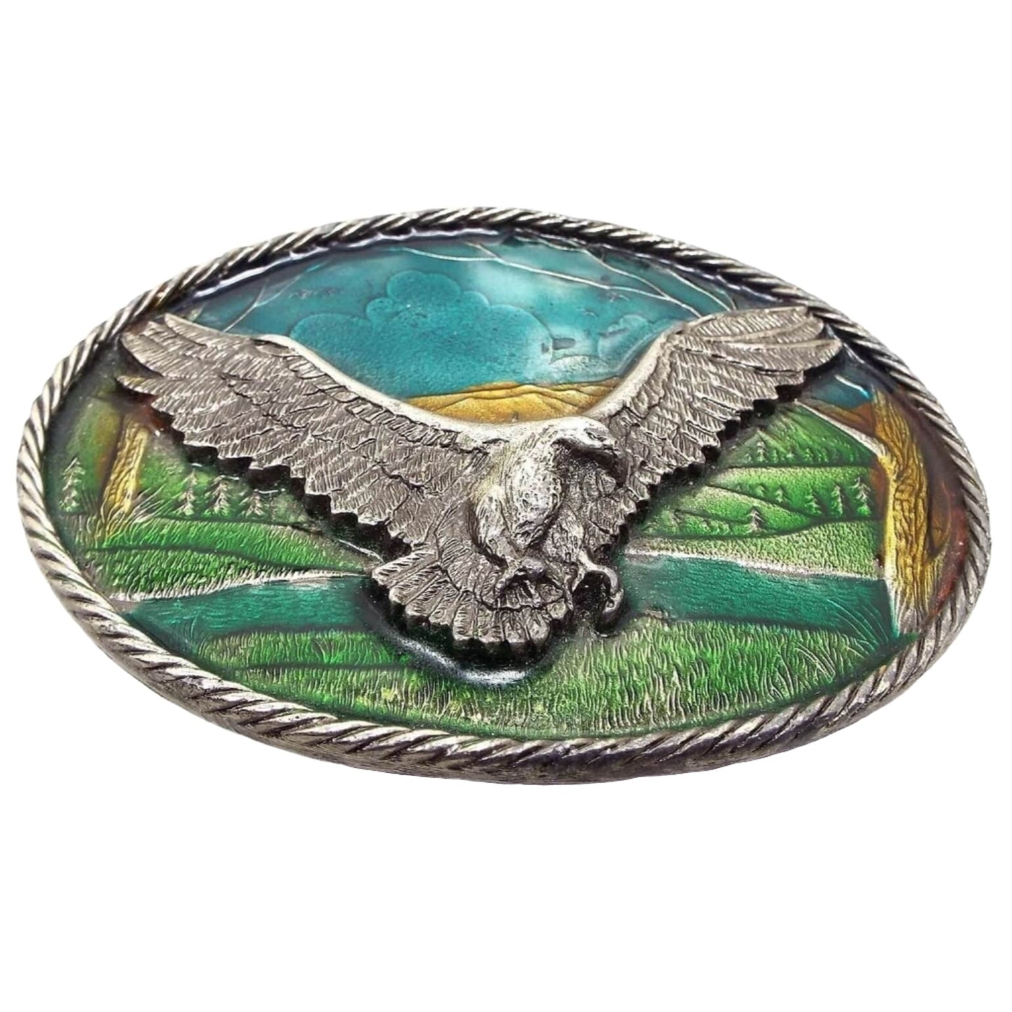 Front view of the retro vintage belt buckle from The Great American Buckle Company. The buckle is a gray pewter oval with braided edge. The design is detailed and textured with a flying eagle with its wings spread in the middle. The background is an enameled scenery of grass, trees, and sky, in shades of blue, green, and brown. 
