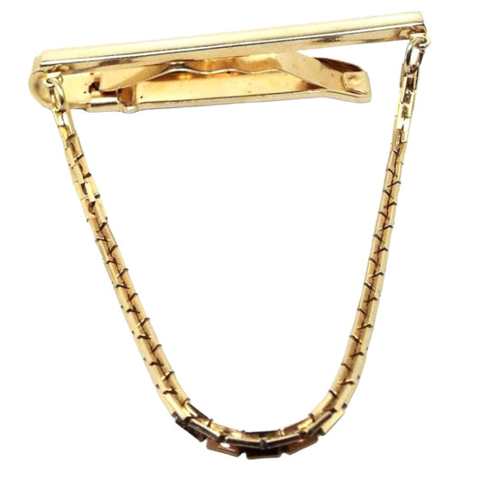 Front view of the retro vintage tie chain bar. The metal is gold tone in color. There is a squared bar on the front and a curved flatter style bar on the back. There is a wavy band of metal in between them to help hold it onto the tie. Square link chain hangs down from each side.