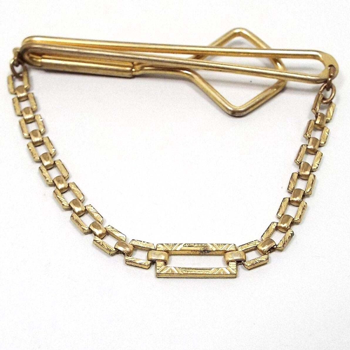 Front view of the 1930's vintage Swank tie chain bar. The metal is gold tone in color. The top bar is an wire open oval that curves around to the back with another wider open oval on the end. A flat style textured square link chain hangs down from each side to a larger textured rectangle link on the bottom.