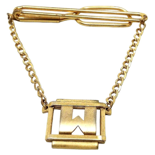 Front view of the 1930's vintage Swank initial tie chain bar. The metal is gold tone in color. The top has a long open oval wire style bar that curves around to the back with a wider open oval at the end. There is curb chain hanging from each end down to an open rectangle charm with a metal bar on top and below it. There is a block letter style initial W in the middle. 