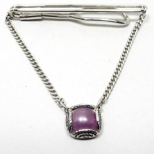 Front view of the Mid Century vintage Swank tie chain bar. The metal is silver tone in color. The top bar is an wire open oval that curves around to the back with another wider open oval on the end. Small curb chain hangs from either side down to a square charm with rounded corners. There is a round domed purple lucite cab in the middle held by 4 large half circle prong areas that has a rope style texture to them. 