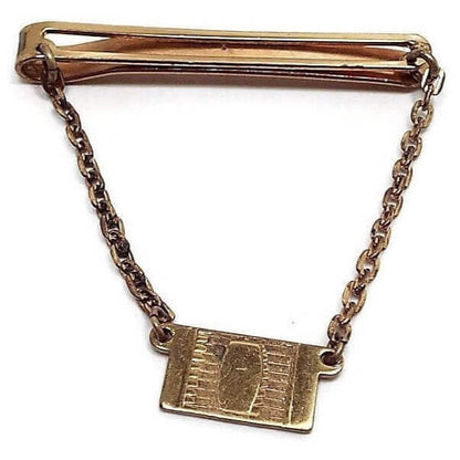 Front view of the 1940's Mid Century Vintage Giant Grip Klick it Dirigold tie chain bar. It is a darkened gold tone, almost brass in color. The top bar is a long open rectangle with rounded ends. Oval link rolo chain hangs done from each side of the top bar down to a rectangle charm. Charm has a modernist design of lines and a rectangle with curved sides in the middle of it.
