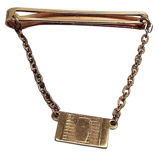 Front view of the 1940's Mid Century Vintage Giant Grip Klick it Dirigold tie chain bar. It is a darkened gold tone, almost brass in color. The top bar is a long open rectangle with rounded ends. Oval link rolo chain hangs done from each side of the top bar down to a rectangle charm. Charm has a modernist design of lines and a rectangle with curved sides in the middle of it.