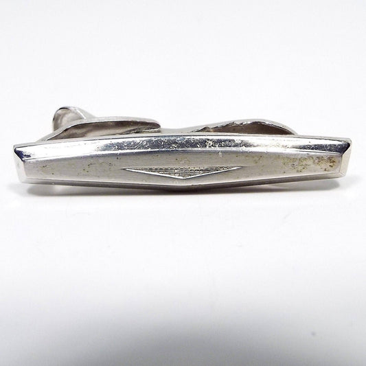 Front view of the Mid Century vintage Hickok tie clip. The metal is silver tone in color. It has a modernist style design with a rectangle shape that has a bowed out middle. At the bottom middle area of the tie clip is an upside down triangle filled with tiny little squares. Part of the alligator style clip on the back is showing.