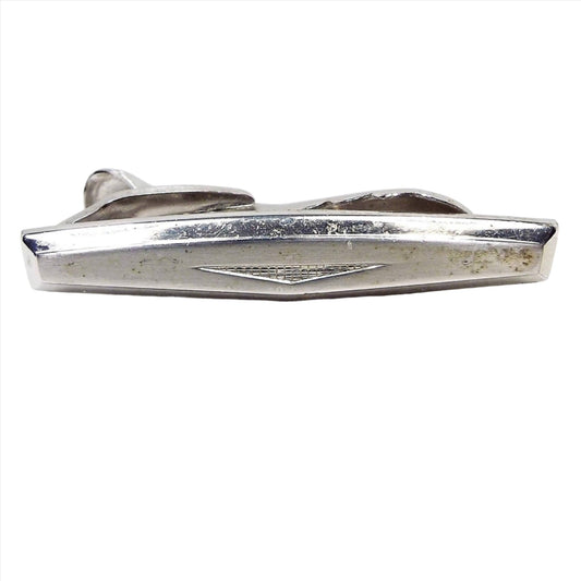 Front view of the Mid Century vintage Hickok tie clip. The metal is silver tone in color. It has a modernist style design with a rectangle shape that has a bowed out middle. At the bottom middle area of the tie clip is an upside down triangle filled with tiny little squares. Part of the alligator style clip on the back is showing.