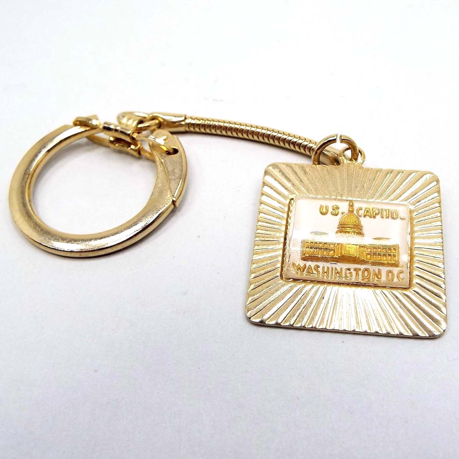 Front view of the Mid Century vintage keychain. The metal is gold tone in color. One end has a latched ring to open and put your keys on. There is a snake chain down to the other end that is square shaped with a plastic cab in the middle. The plastic is clear and shows a raised metallic gold design of the Capitol Building and wording that says "US Capitol Washington DC." The outer edge has textured lines going outward at an angle.