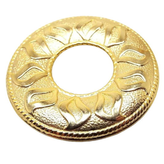 Front view of the retro vintage Kathie Lee scarf clip. It is round in shape with an open middle. There are curvy petal shapes around the open hole to look like a sunflower. The raised petal areas are shiny gold tone in color and the lightly textured background is matte gold tone in color. The edge has a raised rope like design all the way around it.