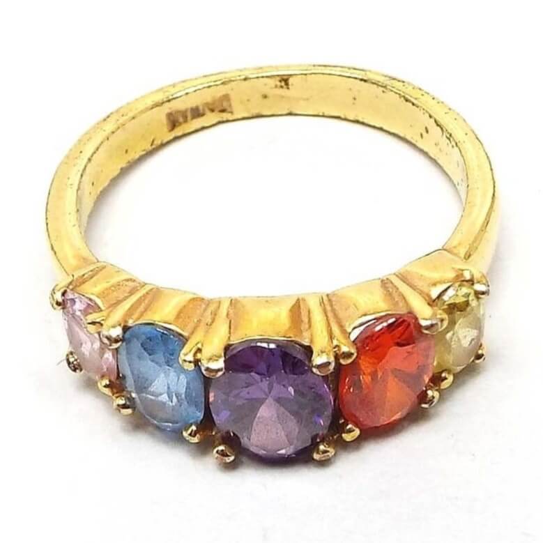 Angled front and side view of the retro vintage rainbow style rhinestone ring. The metal is gold tone in color. There are five multi color oval prong set rhinestones on top in a row that are graduating in size with the largest being in the middle. The colors are light pink, light blue, purple, orange, and light yellow. Taiwan is stamped on the inside of the band.