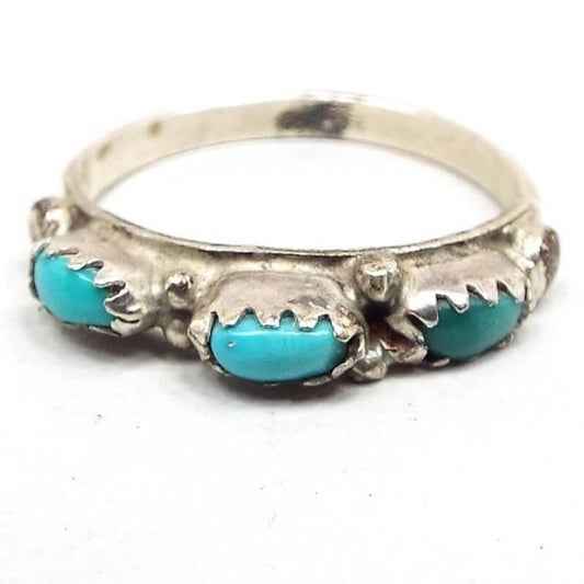 Angled front and side view of the retro vintage faux turquoise ring. The top has three small raised oval bezels with prong like sides holding resin imitation turquoise cabs in a puffed oval shape. Setting and band are silver tone in color. There is a dark spot on the silver color on the inside of the band on the bottom.