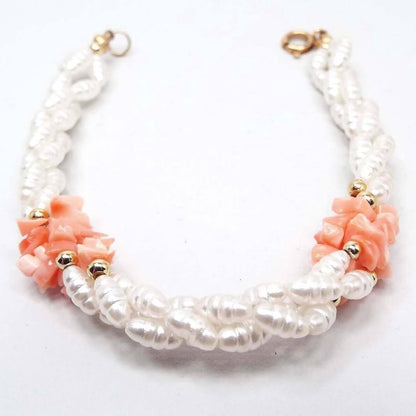 Side view of the retro vintage faux pearl and shell beaded bracelet. The beaded strands are twisted and braided down the length of the bracelet. strands are beaded with coated glass rice shaped imitation pearl beads in white. On each side of the bracelet towards the top are larger chip style shell beads in a salmon pink color. There are some small round gold tone beads by the shell beads. The end of the bracelet has a gold tone round spring ring clasp. 