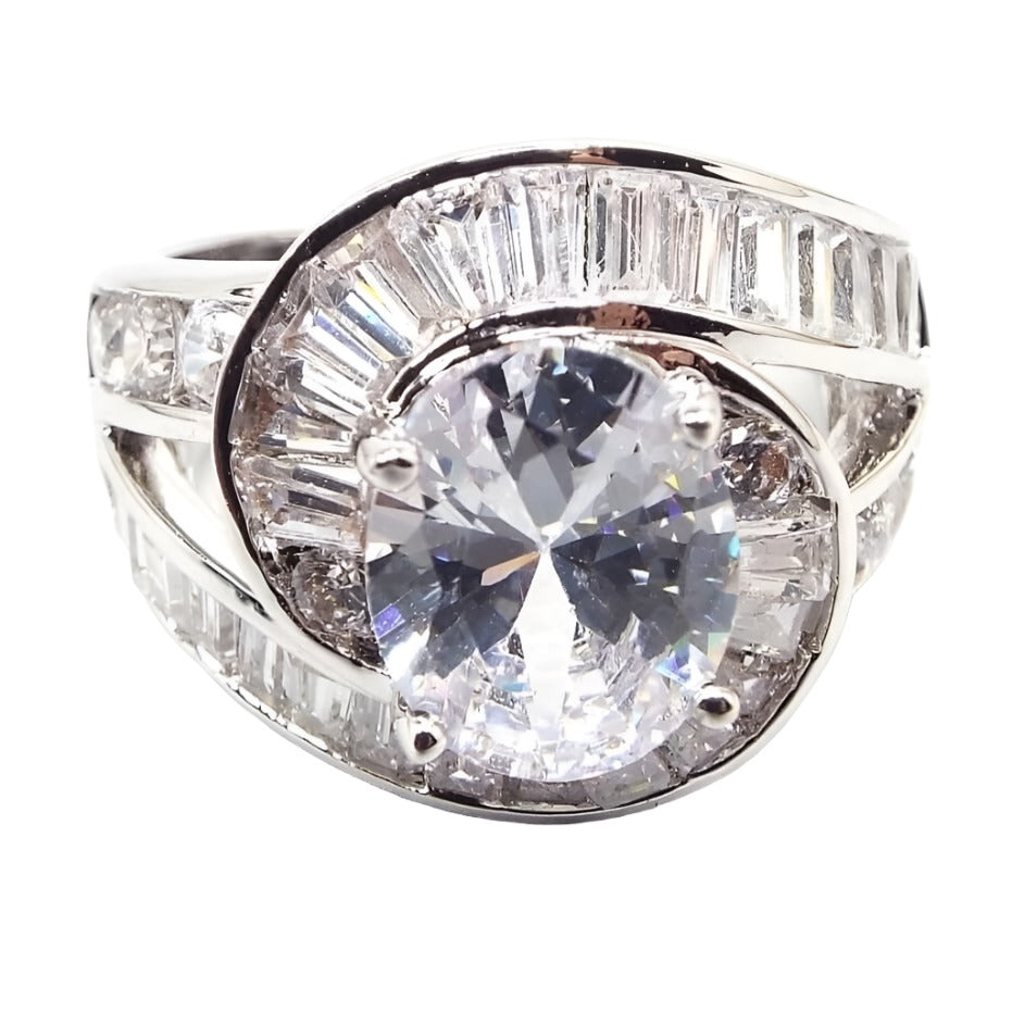 Front view of the retro vintage glitzy glam rhinestone cocktail ring. It is gold filled but is silver in color. The top of the ring has a larger sized round rhinestone. The band is split on either side and one row has small round rhinestes and the other row has baguette and trapezoid shaped rhinestones. The row with the baguettes and trapezoids bypasses and curves around either side of the middle rhinestone. 
