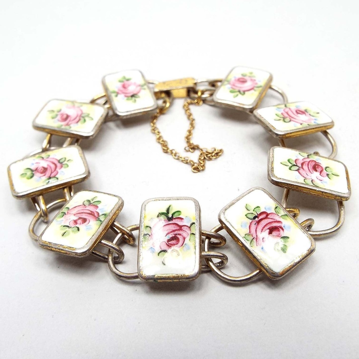 Top view of the Mid Century vintage Bliss Brothers floral bracelet. The snap lock clasp, safety chain, and some areas of the rectangle links are vermeil gold plated over sterling. The rest is silver in color. There is wear the the plating seen around the edges of the links and clasp. The porcelain cabs are white with pink roses with green leaves painted on them.