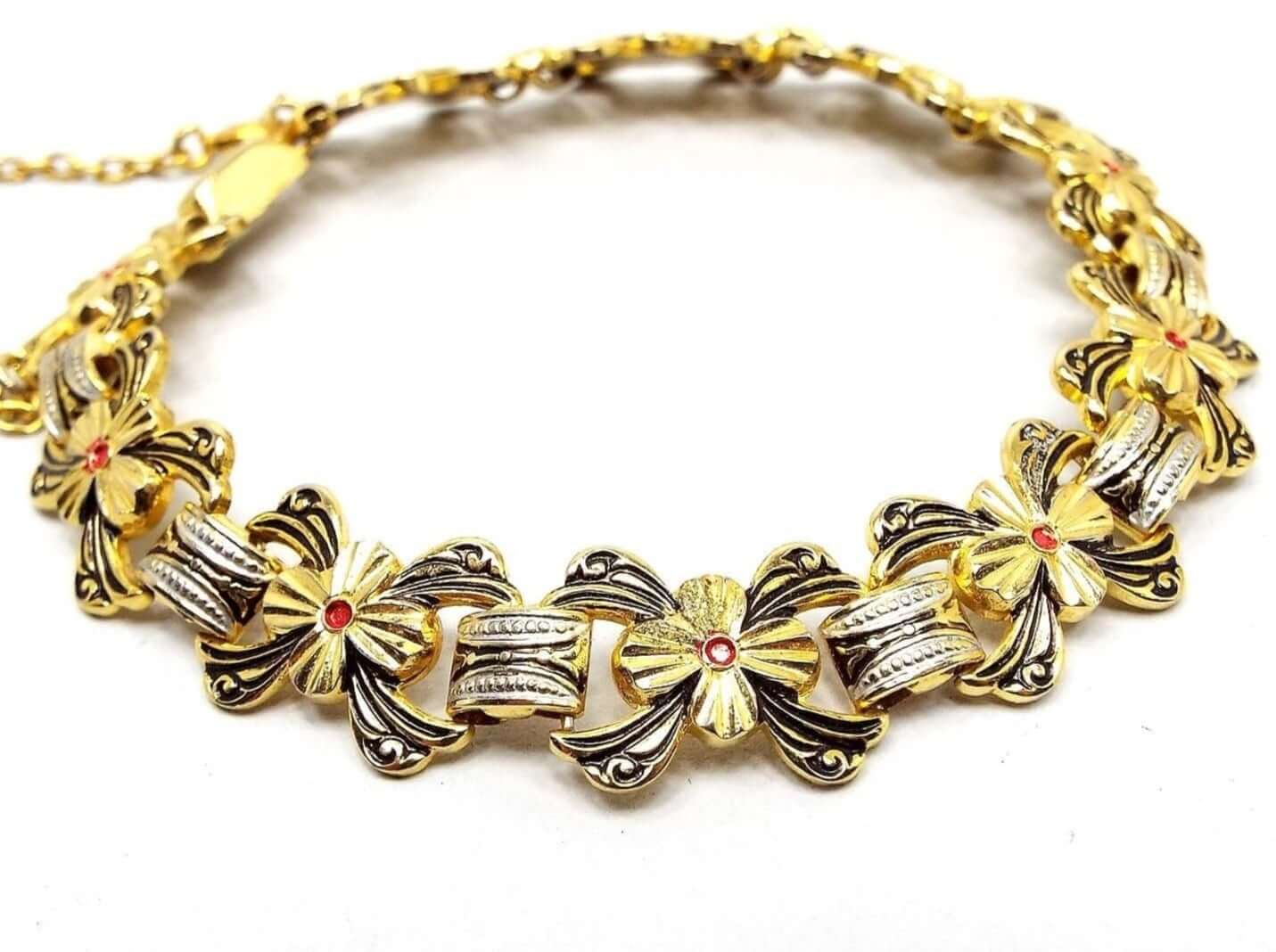 Top view of the retro vintage Damascene style link bracelet. Each link has a bow like design with textured gold tone that has black in the indented areas. In the middle is a flower with 4 petals and a red dot in the middle. Those links are connected by domed rectangle links with a dot pattern. The domed links have silver tone color metal on the sides and the rest is gold tone. At the end of the bracelet is a box clasp and a thin safety chain near the clasp area that has a round spring ring clasp.