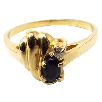 Angled front view of the retro vintage sapphire and rhinestone ring. The metal is gold tone in color. On the top of the band is a scalloped half shell shape design. On the side of that is an oval prong set black sapphire that has just a tiny hint of blue when magnified under bright light settings. Next to the sapphire is a tiny round prong set rhinestone. 
