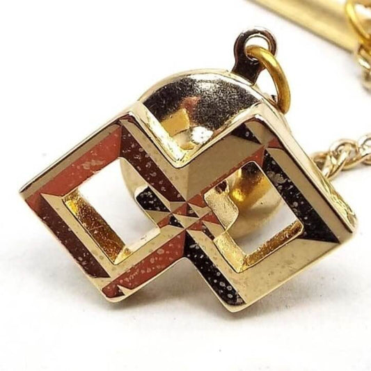 Front view of the retro vintage tie tack. There are two squares side by side in a diamond shape and the inside corners are overlapping. Each one is faceted and slopes slightly inward. The middle area of each is open. The metal is shiny gold tone in color, but there are some tiny lighter spots from age when magnified. There is a round clutch on the back that has a chain with a small bar on the end.