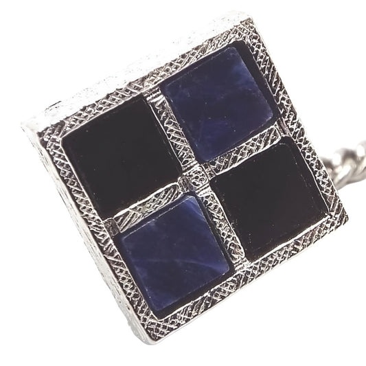 Front view of the Mid Century vintage Dante tie tack. Tie tack has textured silver tone color with a square shape. There are four squares of gemstone cabs on the front. Two are black onyx and the other two are dark blue sodalite gemstone. The gemstone types are diagonal to one another. There is a chain on the back clutch with a small bar at the end. 