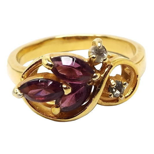 Angled front view of the retro vintage rhinestone cocktail ring. The metal is gold tone in color. On the top is a large sideways curled S shape. There is a small round prong set clear rhinestone on the inside and outside edge of the smaller curve. The larger curve has three marquis shape purple rhinestones that are angled in a leaf like design. 
