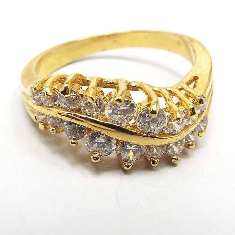 Angled top and side view of the retro vintage cubic zirconia cocktail ring. The top has a curved wavy design. There are two rows of CZ stones that are graduated in size from largest in the middle to smaller at the ends. All are prong set and sparkling clear in color. The setting and curvy metal band between the stones is plated gold tone in color. 