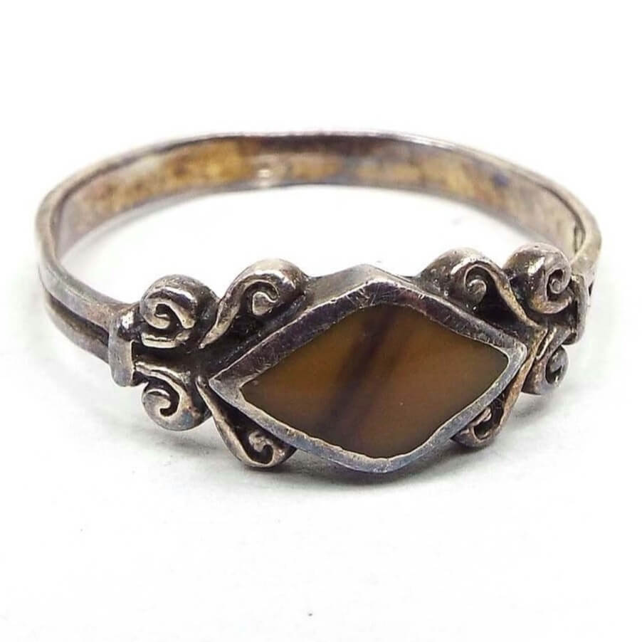 Angled front view of the retro vintage Nelvin Chee ring. the sterling silver has darkened from age to a light gray in color. The top of the ring has a diamond shaped bezel with an inlaid unidentified gemstone in it. The gemstone has diagonal stripe variations of color from mustard yellow to brown. On either side of the gemstone is a curled scroll like design and then the band is flatter style with a line in the middle all the way around it.