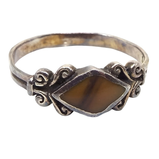 Angled front view of the retro vintage Nelvin Chee ring. the sterling silver has darkened from age to a light gray in color. The top of the ring has a diamond shaped bezel with an inlaid unidentified gemstone in it. The gemstone has diagonal stripe variations of color from mustard yellow to brown. On either side of the gemstone is a curled scroll like design and then the band is flatter style with a line in the middle all the way around it.