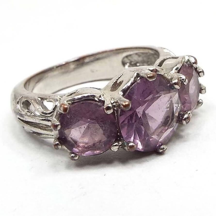 Angled front and side view of the retro vintage purple multi stone ring. The top has three round larger sized purple rhinestones. The one in the middle is larger than the other two. All are prong set. The metal is silver tone in color. The sides of the band near the stones has a cut out style design.