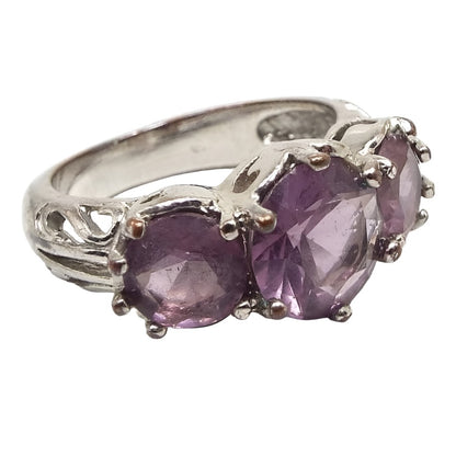 Angled front and side view of the retro vintage purple multi stone ring. The top has three round larger sized purple rhinestones. The one in the middle is larger than the other two. All are prong set. The metal is silver tone in color. The sides of the band near the stones has a cut out style design.