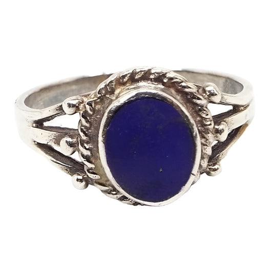 Angled front and side view of the retro vintage sterling silver Southwestern style gemstone ring. The top middle has a flat oval bezel set lapis lazule cab in a shade of darker blue. There is a rope style design around the edge of the bezel. The band splits into three parts on either side of the gemstone and has a rounded dot at the end of each. 