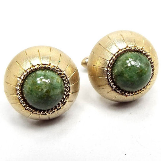 Angled front view of the Mid Century vintage Swank gemstone cufflinks. The fronts are round domed with bezel set round domed serpentine cabs in the middle that are a mossy green in color. The other edge of the cufflinks has an etched line design running from front to back. The metal is gold tone in color.