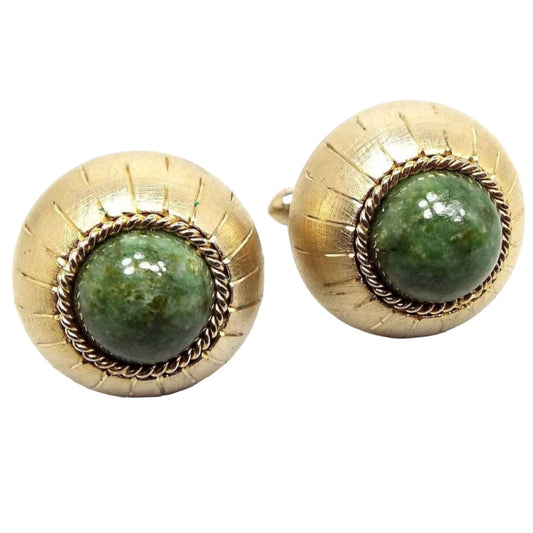Angled front view of the Mid Century vintage Swank gemstone cufflinks. The fronts are round domed with bezel set round domed serpentine cabs in the middle that are a mossy green in color. The other edge of the cufflinks has an etched line design running from front to back. The metal is gold tone in color.